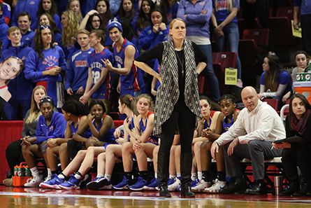 Girls Basketball coach stands infront of players on the team bench watching the game. 